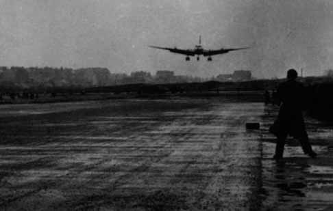Aircraft landing on a wet runway at dusk with a figure watching from the edge