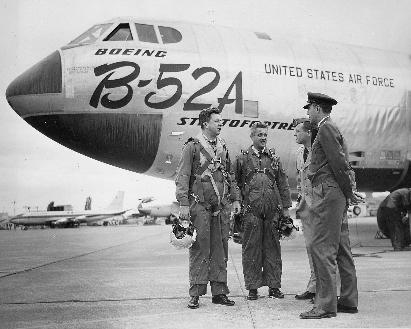 Two flight-suited airmen and two officers stand beneath the nose of a Boeing B-52A Stratofortress on the factory ramp