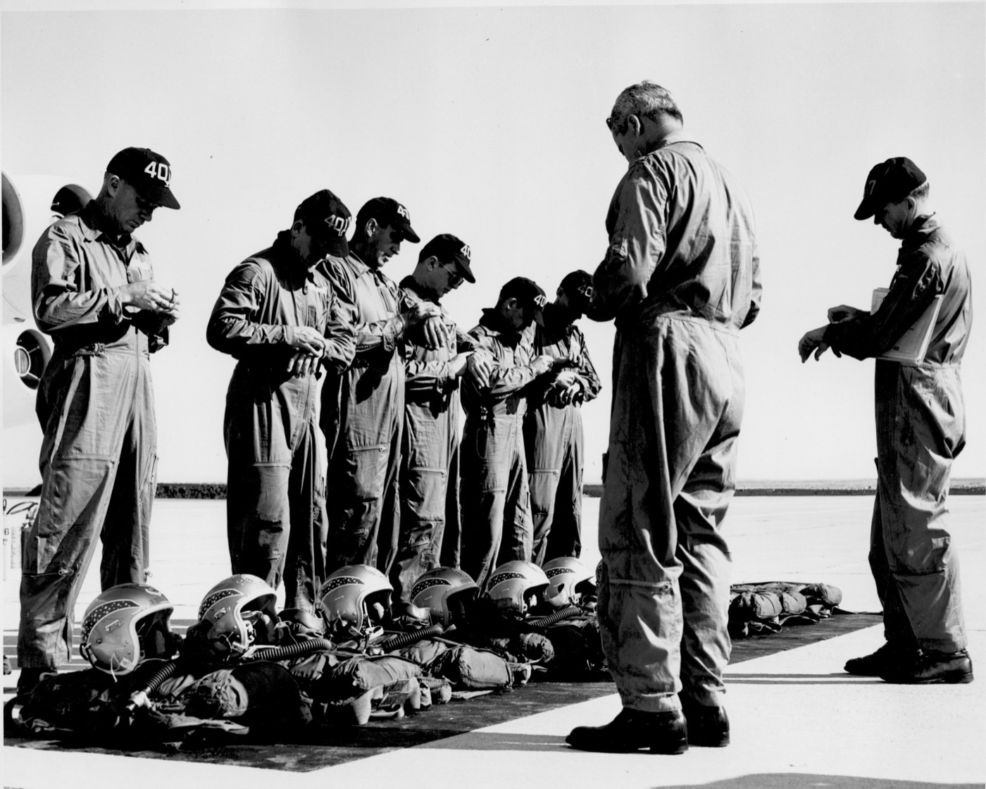 A line of B-52 aircrew in flight suits check their gear during a pre-flight, flight helmets laid out on the ramp, an officer facing the line