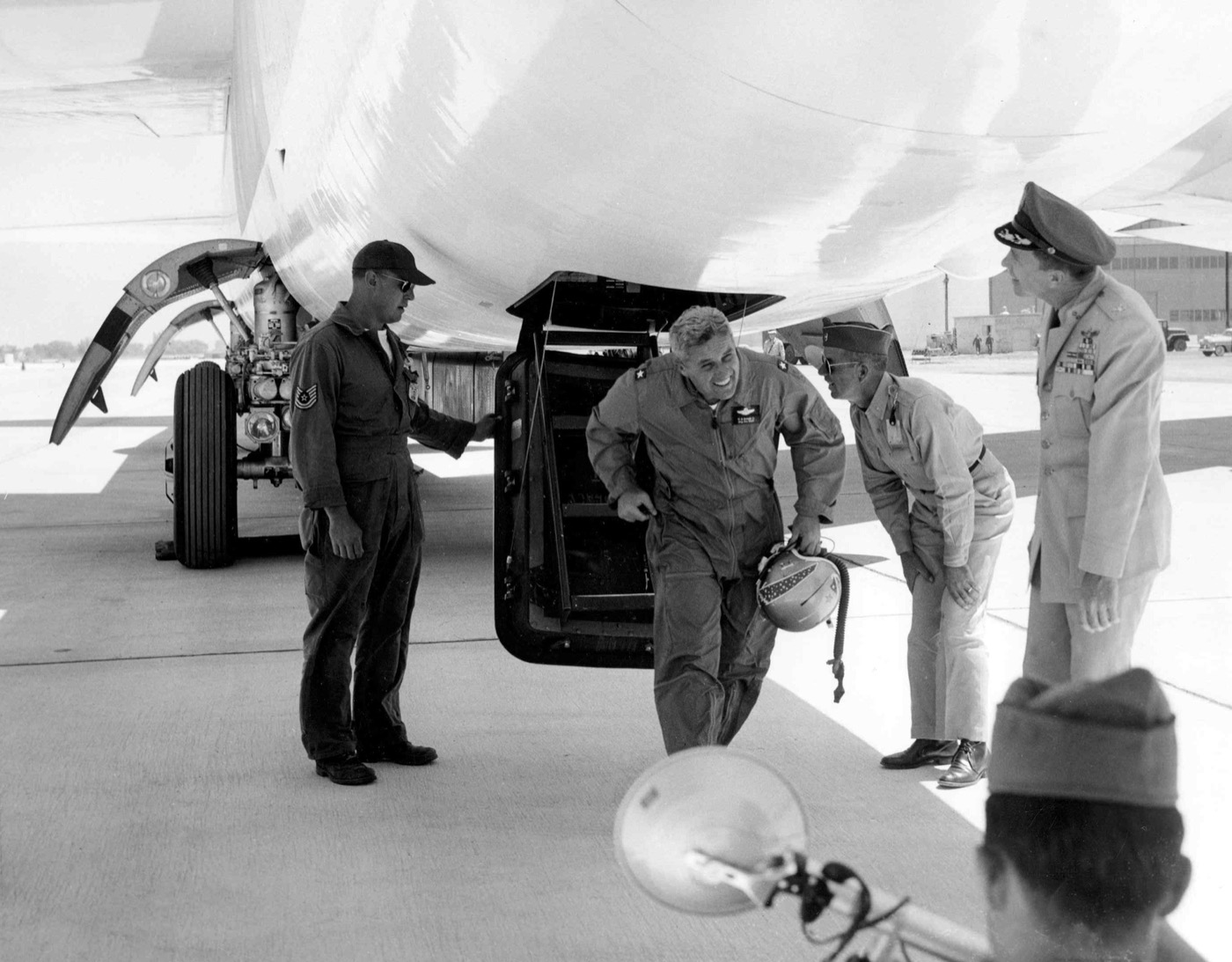 A smiling officer in a flight suit steps out of the belly hatch of a B-52, greeted by officers and a crew chief