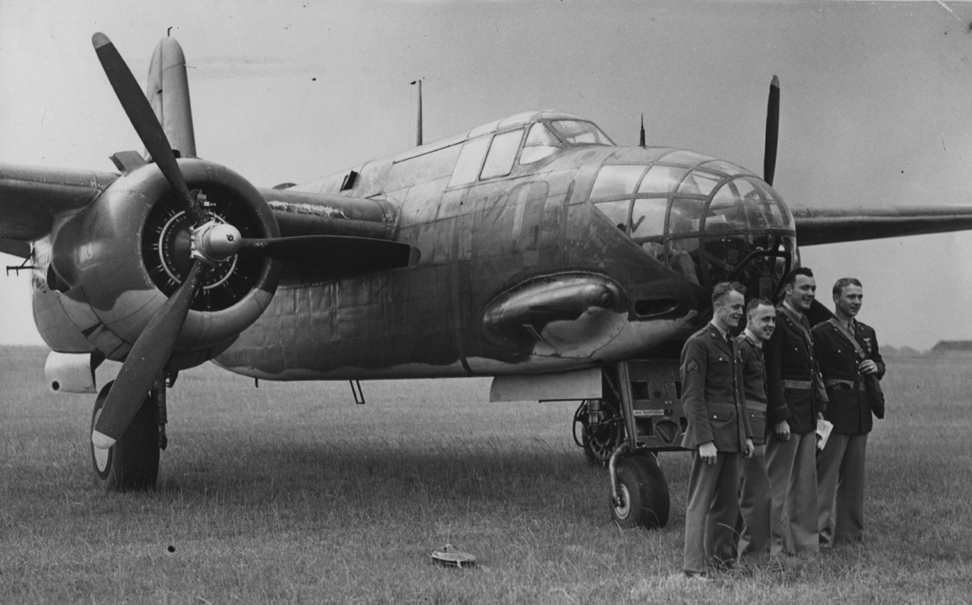 Four airmen in service uniform stand in a row at the glazed nose of a twin-engine Douglas Boston III light bomber parked on a grass airfield; the two at left wear enlisted stripes, the two at right are officers.