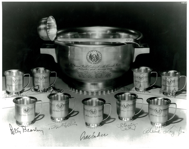 A sterling silver twin-handled punch bowl with a ladle resting on its rim, engraved with a winged emblem and dedication text, surrounded by nine matching engraved cups arranged in two rows; ink signatures of the honored officers are written across the photograph beneath the set.